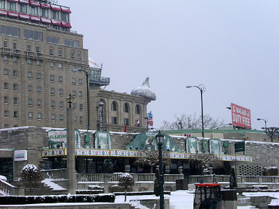 [A tan brick or stone building, sitting beside a much larger building with lots of windows, has a large silver replica of a Hershey candy kiss. There is snow on the kiss as well as on the ground.]